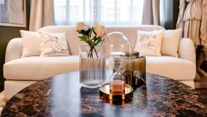 Ett runt bord av marmor med en vas av glas, en genomskinlig behållare av glas och en liten metallbricka med ett ljus. I bakgrunden finns en vit soffa med dekorativa kuddar och en staty.