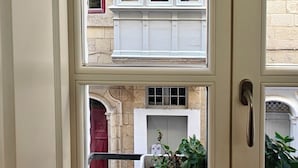 A multi-pane window with a view of a building with red and white doors, a stone wall, and a balcony with plants. - Traditional Maltese 1BR Home - Valletta (Valletta)