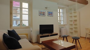 A living room with a sofa, a TV, a coffee table, and a desk with a chair. There are two windows with curtains, a white shelf unit, and a wall-mounted light fixture. - Traditional Maltese 1BR Home - Valletta (Valletta)