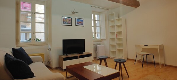 A living room with a sofa, a TV, a coffee table, and a desk with a chair. There are two windows with curtains, a white shelf unit, and a wall-mounted light fixture.
