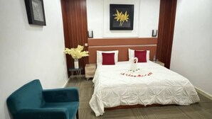 Et hotelværelse med en seng, en blå lænestol, et lille bord med en blomstervase og et billede i ramme på væggen.