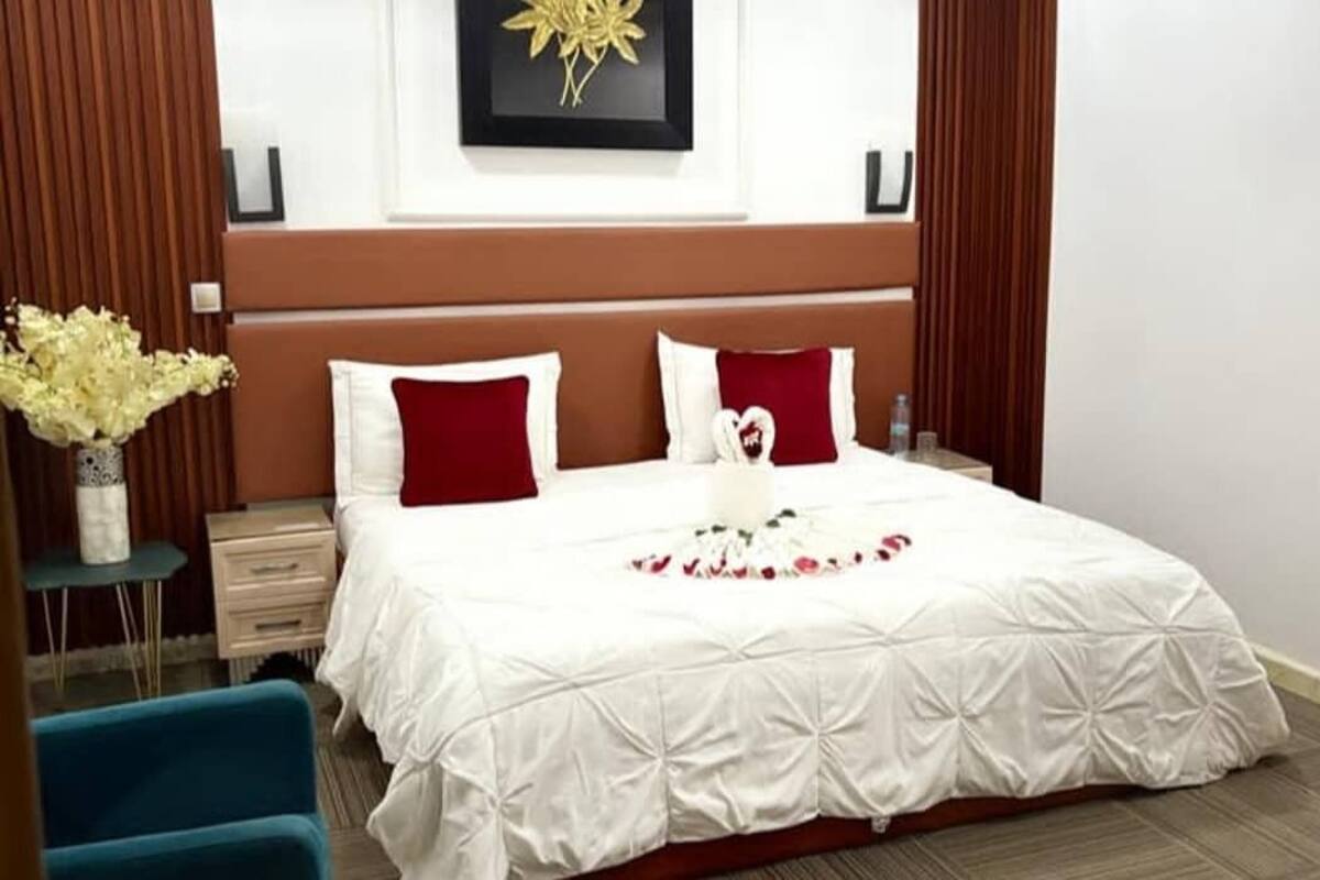 Ein ordentlich bezogenes Bett mit weißer Bettwäsche und roten Kissen, eine Vase mit weißen Blumen und ein gerahmtes Bild an der Wand.