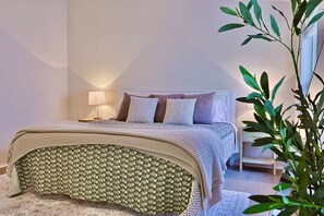 Un lit bien fait, recouvert d’une couvre-lit à motifs floraux et d’une couverture tricotée, une lampe de chevet et une petite table de chevet avec une lampe.
