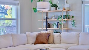 A living room with a white sofa, a side table, and a wooden shelving unit with plants and decorative items. - Happy's Hideaway w/ Hot Tub, Sleeps 12, Downtown! (Ellensburg)