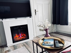Ein Wohnzimmer mit Kamin, einem Wand-Fernseher, einer Vase mit Blumen und einem Tablett mit Buch und Fernbedienung.