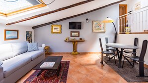Una sala de estar tipo loft con techo alto, vigas a la vista, un sofá, una mesa de centro, una mesa de comedor con sillas y un televisor montado en la pared.