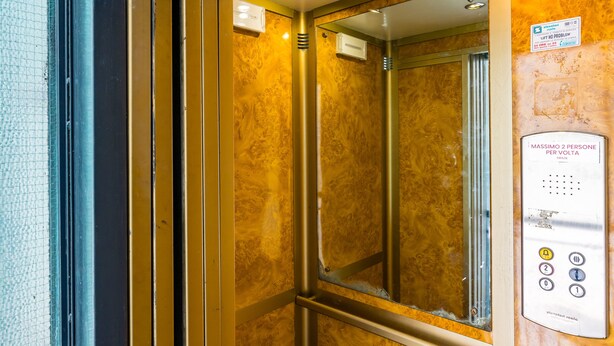 Interior de ascensor con pared espejada, panel de control y puerta metálica.