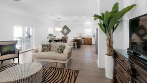 A modern living room with a sofa, coffee table, and a large plant. There is a kitchen in the background with white cabinets and a refrigerator. - Epic! Ocean views! Sleeps 20! Pool! Golf cart! (Destin)