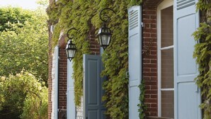 A brick building with ivy-covered walls, a blue door, and two windows with shutters. - RIVE Side House    . (Muids)