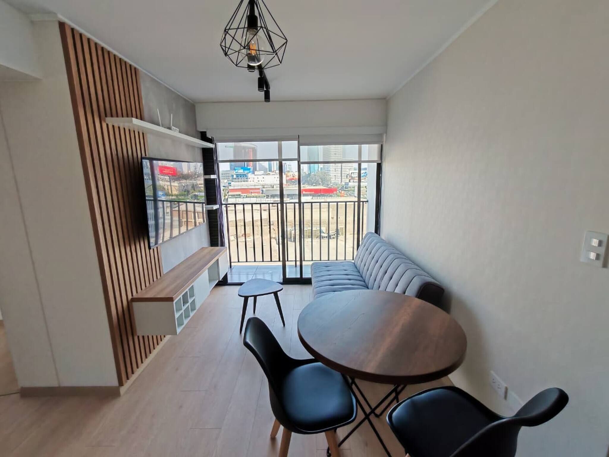 A modern living room with a dining area, a sofa, a TV mounted on a wooden panel, and a balcony with a view of the city.
