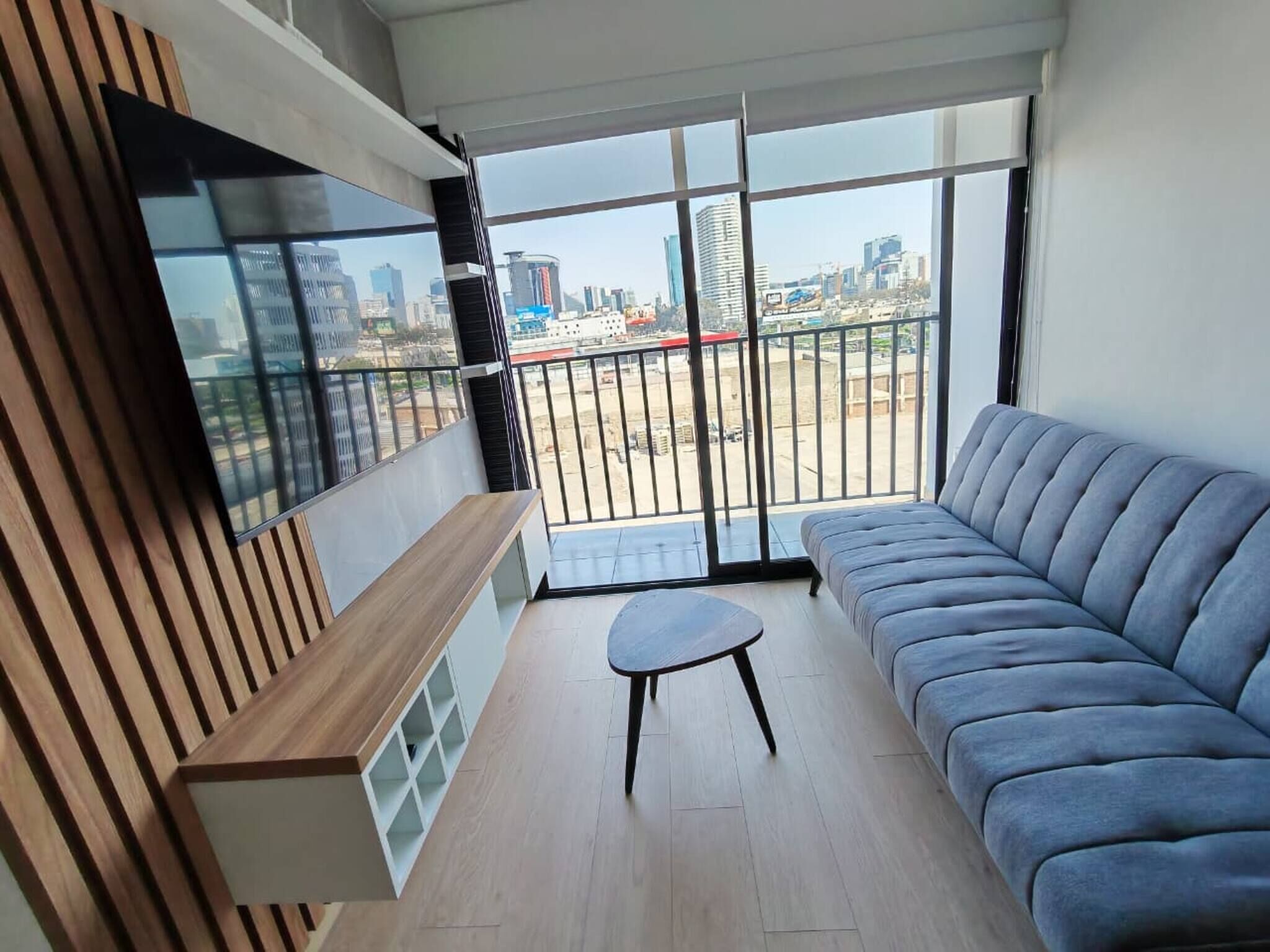 A modern living room with a grey sofa, a small round table, and a wooden bench. A large TV is mounted on the wall, and the room has a view of a cityscape through large windows.