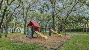 A playground with a red-roofed playhouse and two slides in a park with trees. - Gorgeous 1 BR top floor + Private Beach + pools + hot tub Washer Dryer + Balcony (St. Petersburg)