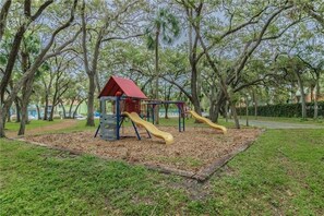 Ein Spielplatz mit einer roten Spielhütte und zwei Rutschen in einem Park mit Bäumen.