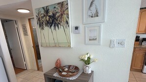 A hallway with a white textured wall, a framed picture of palm trees, a thermostat, and a small shelf with decorative items. - Gorgeous 1 BR top floor + Private Beach + pools + hot tub Washer Dryer + Balcony (St. Petersburg)