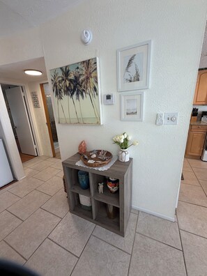 A hallway with a white textured wall, a framed picture of palm trees, a thermostat, and a small shelf with decorative items. - Gorgeous 1 BR top floor + Private Beach + pools + hot tub Washer Dryer + Balcony (St. Petersburg)