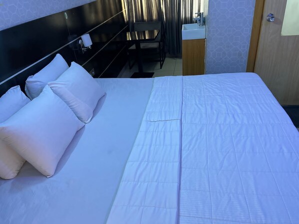 A neatly made bed with white bedding and pillows, a wooden headboard, a small table with a chair, and a bathroom with a sink and mirror. - Plot1 Suites (Ibadan)
