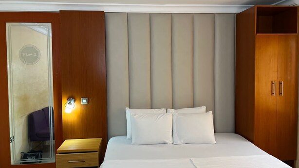 A hotel room with a bed, a wooden wardrobe, a mirror, and a small table with a lamp.