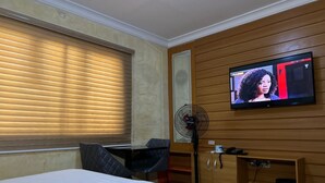A hotel room with a bed, a desk, a chair, a TV mounted on the wall, and a standing fan.
