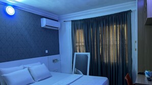 A hotel room with a bed, a desk, an air conditioner, a window with curtains, and a nightstand.