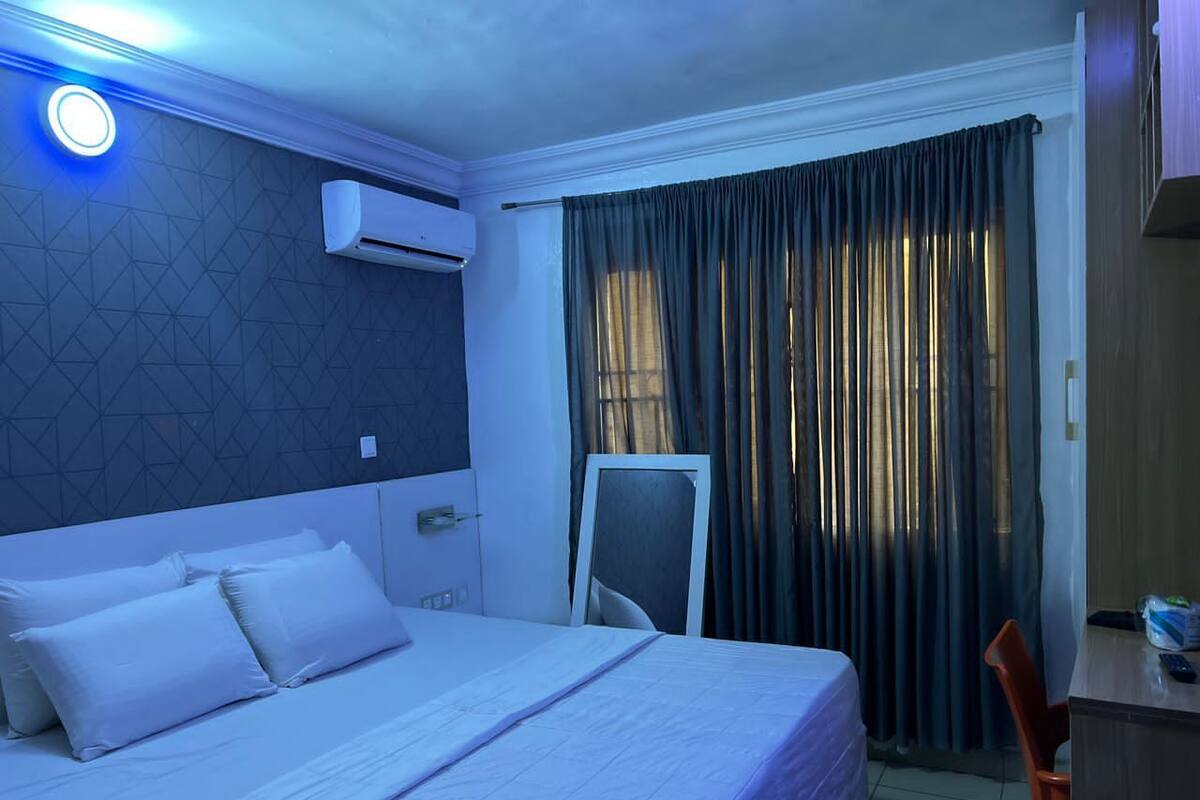 A hotel room with a bed, a desk, an air conditioner, a window with curtains, and a nightstand.