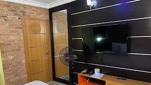 A bedroom with a bed, a TV mounted on the wall, a wooden wardrobe, and a fan.