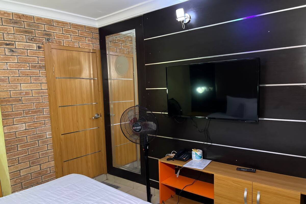 A bedroom with a bed, a TV mounted on the wall, a wooden wardrobe, and a fan.