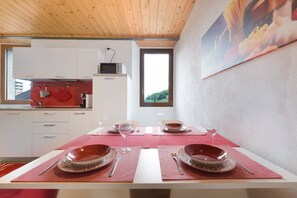 A dining area with a table set for two, a kitchen in the background, and a window with a view.