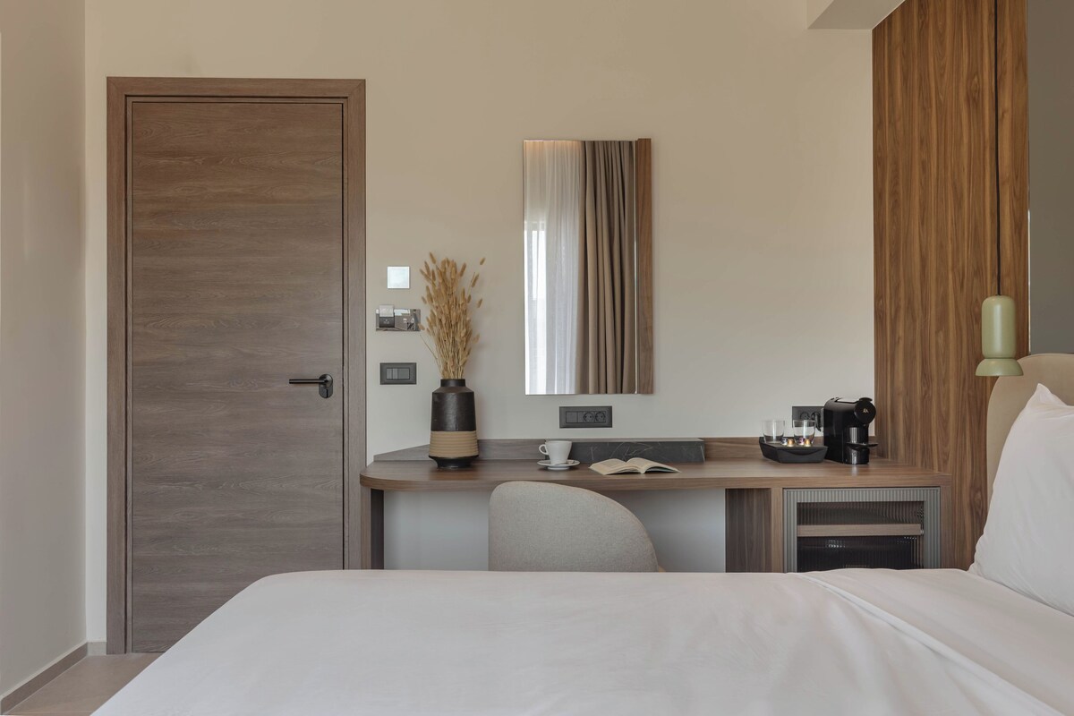 A modern hotel room with a wooden door, a desk with a vase, a mirror, a bed, and a lamp.