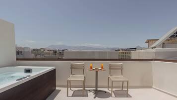A rooftop terrace with a hot tub, two chairs, and a table with a plate and drinks.