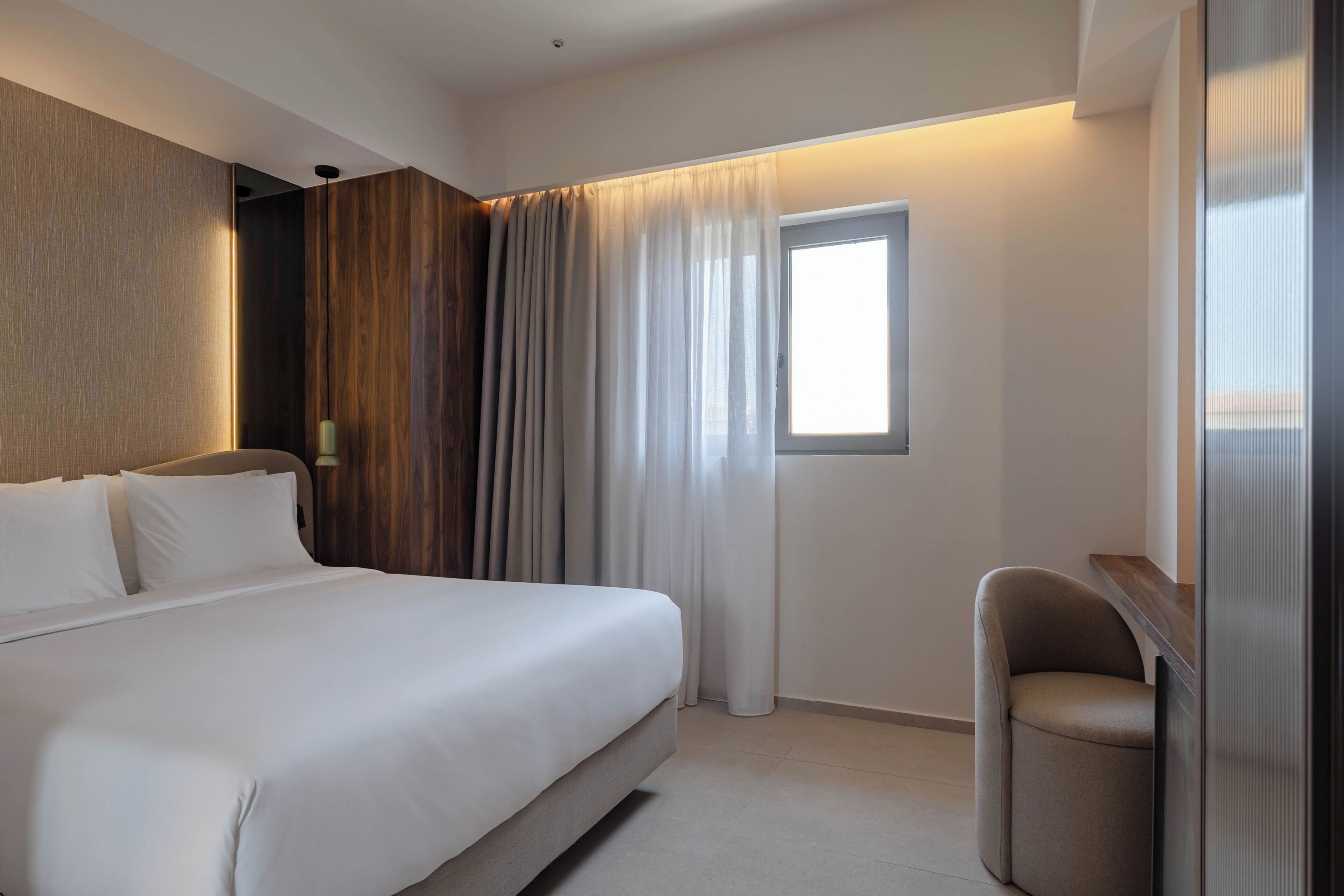 A hotel room with a bed, a chair, a window with curtains, and a wall with a wooden panel.