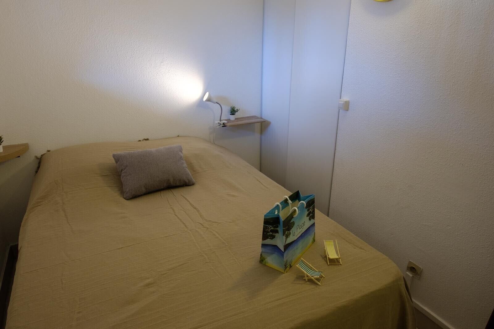 Ett sovrum med en säng, en kudde, ett nattduksbord, en lampa, en hylla med en växt och en väska med toalettartiklar.