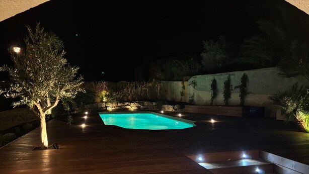 A nighttime view of a swimming pool with a wooden deck and steps leading to it, surrounded by plants and a stone wall. - Villa with pool 2.5km from Saint-Tropez (Gassin)