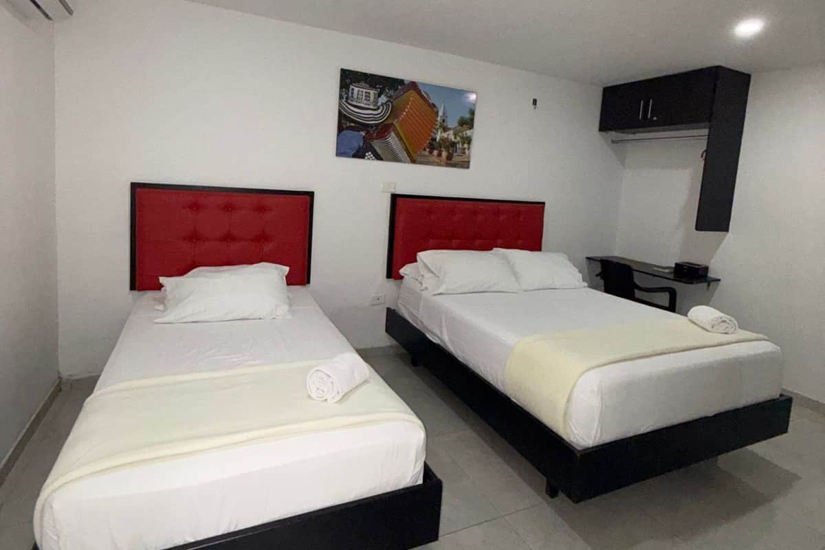 A hotel room with two beds, each with a red headboard, white bedding, and a small table with a lamp. There is a picture on the wall and a black cabinet above the beds.