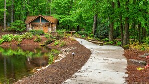 A cabin with a stone foundation and wooden exterior, surrounded by a forest and a pond. - Best of Bear Lake | Pool • Golf • Lake & Mtn Views (Tuckasegee)