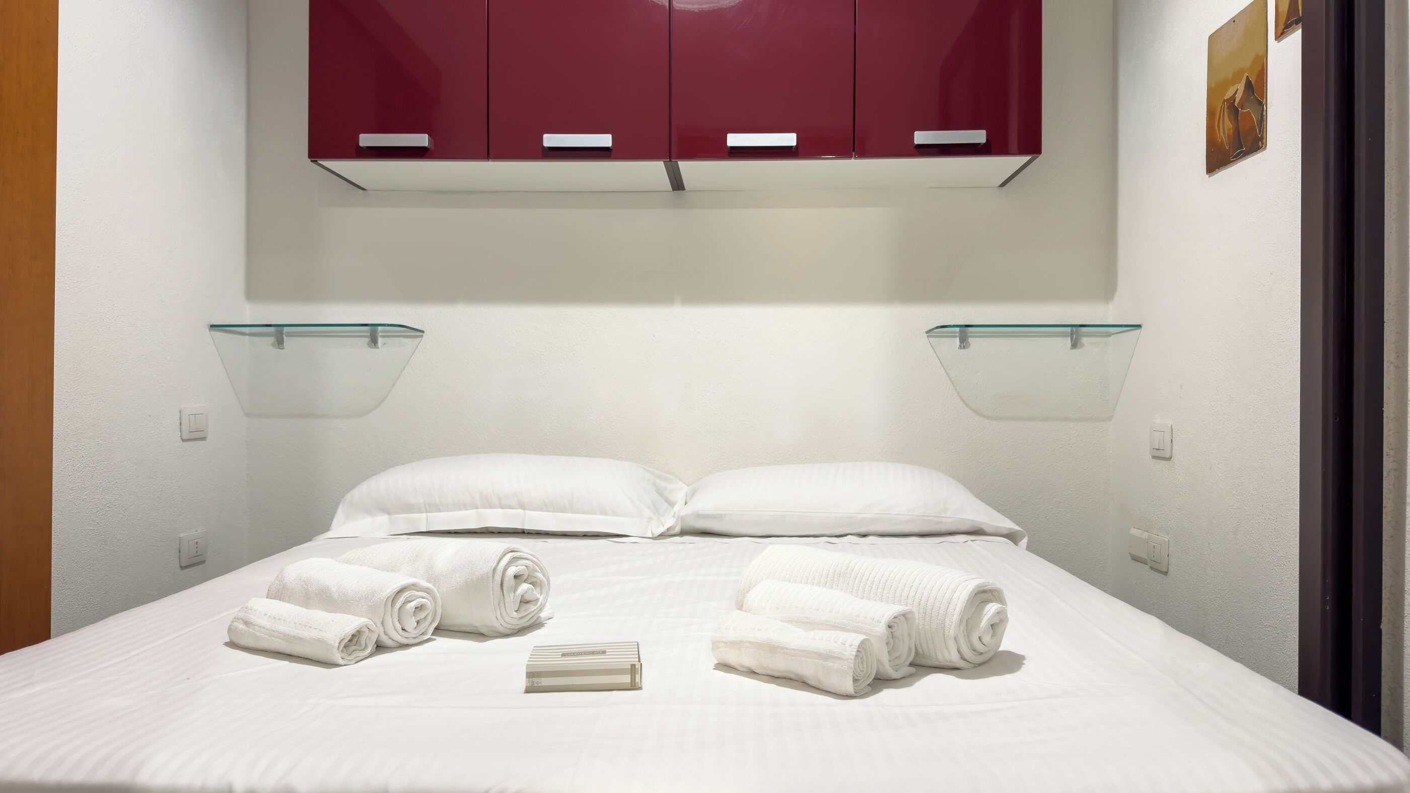 A hotel room with a bed, white linens, a red wardrobe, and glass shelves.