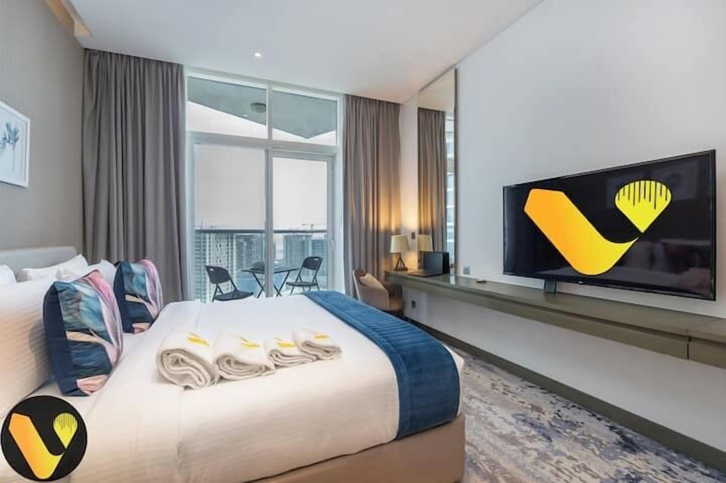 Quarto de hotel com cama, escrivaninha, televisão e janela com cortinas.