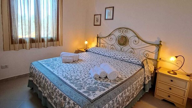 A neatly made bed with a patterned bedspread and pillows, a bedside table with a lamp, and a window with curtains. - Residenze Cielo e Mare - Sole (San Teodoro)