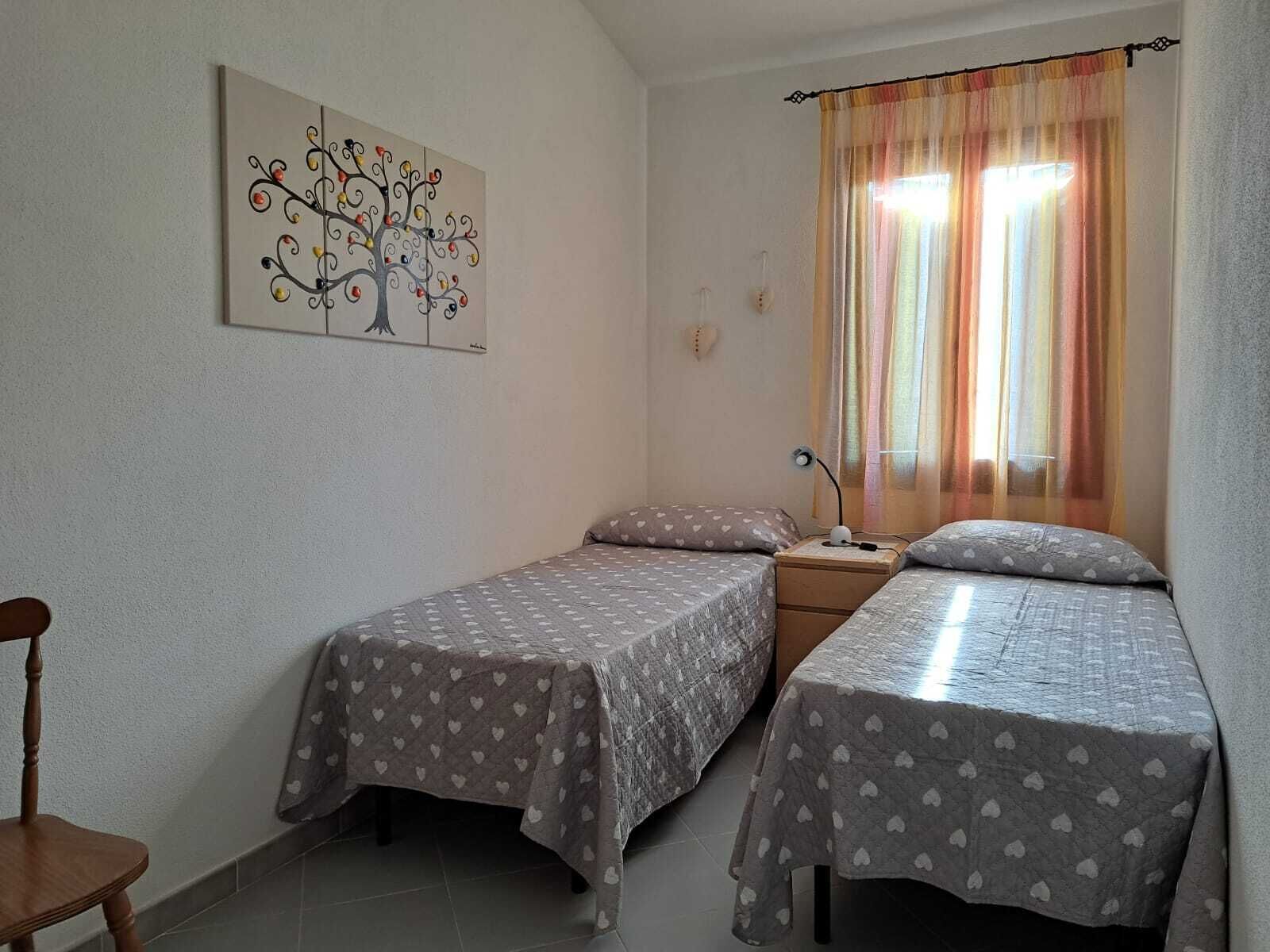 A small, cozy room with two single beds, a wooden chair, a bedside table with a lamp, and a wall-mounted painting.