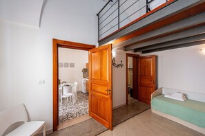 Une petite pièce comprenant un lit, une petite table et une chaise. Une porte donne accès à une autre pièce où se trouvent une table à manger et des chaises.
