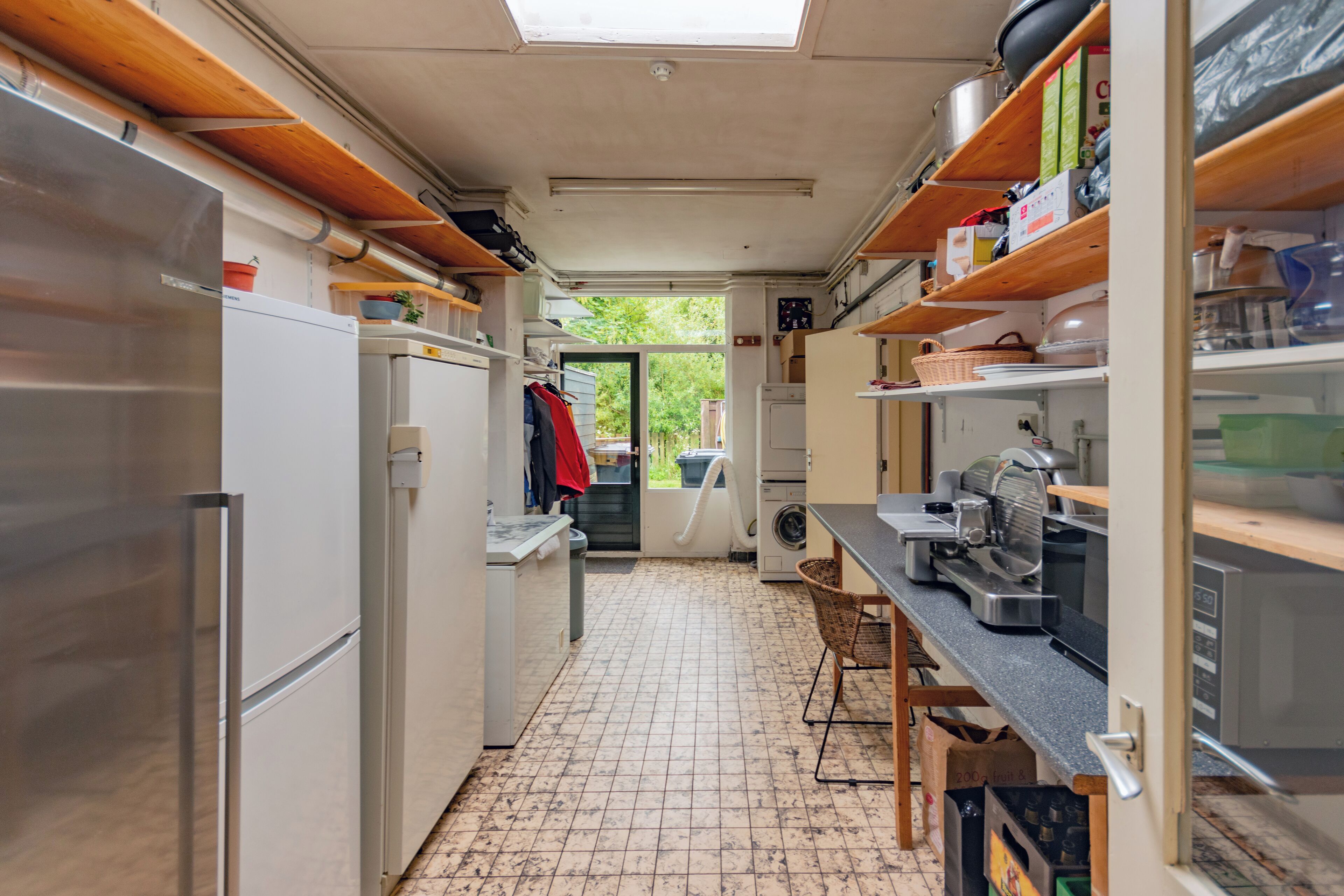 Eine kleine Küche mit Fliesenboden, einem Einbauofen und einem Spülbecken. Über der Arbeitsplatte befinden sich Holzregale, und ein Fenster sorgt für natürliches Licht.