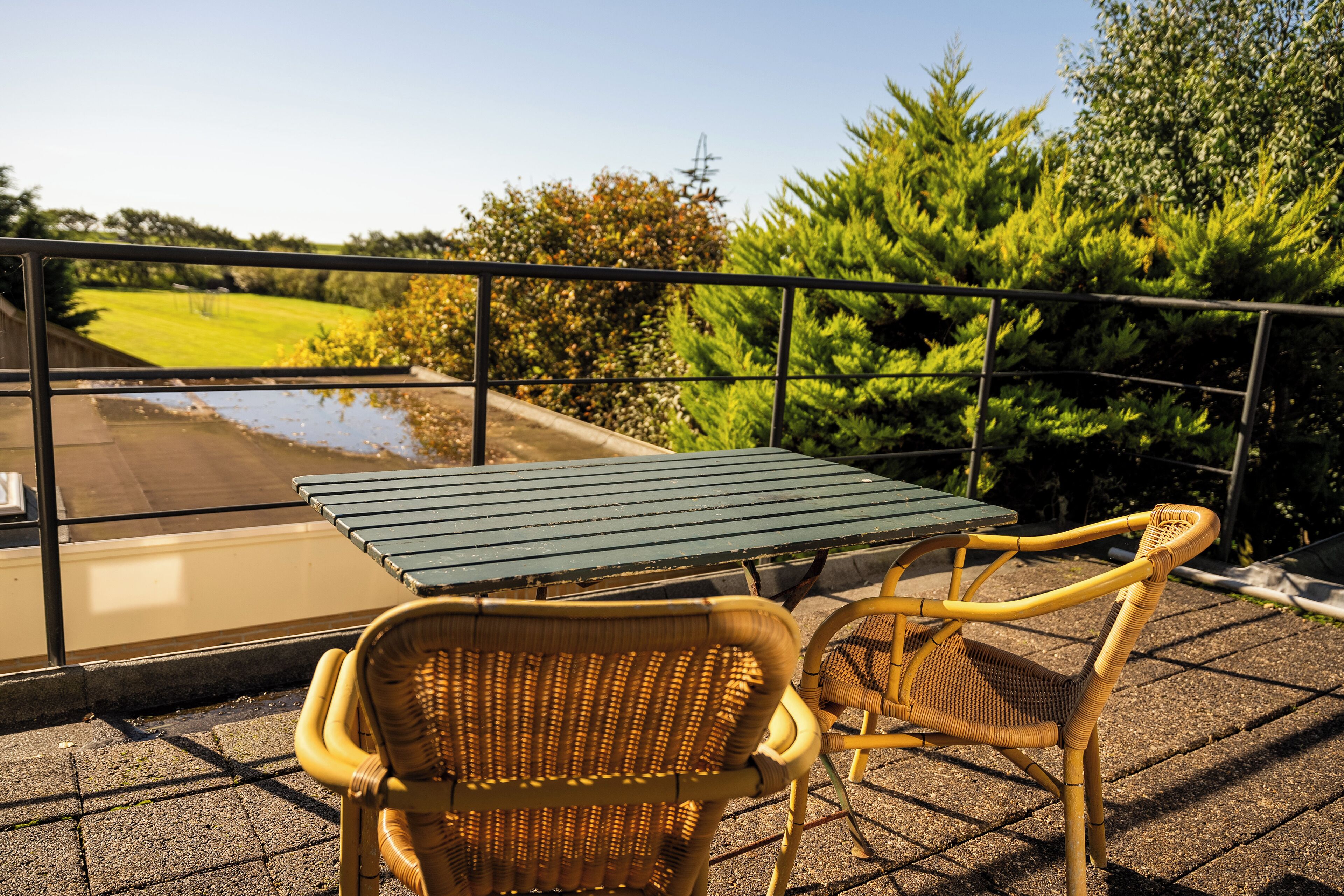 A balcony with a table and two wicker chairs, overlooking a garden and a pond.