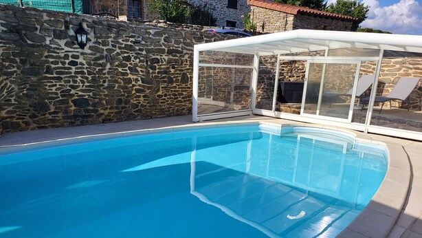 A clear blue swimming pool with a stone wall and a glass-enclosed lounge area. - Au bon accueil gîte with covered and fenced swimming pool, garden, hiking trails (Bonnac)