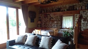 A cozy living room with a stone wall, a wooden ceiling, a black leather sofa with cushions, a small table, and a window with curtains. - Au bon accueil gîte with covered and fenced swimming pool, garden, hiking trails (Bonnac)