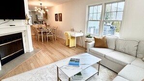 Una sala de estar moderna con un sofá, una mesa de centro y una chimenea. Hay un comedor con una mesa y sillas, y una cocina al fondo.