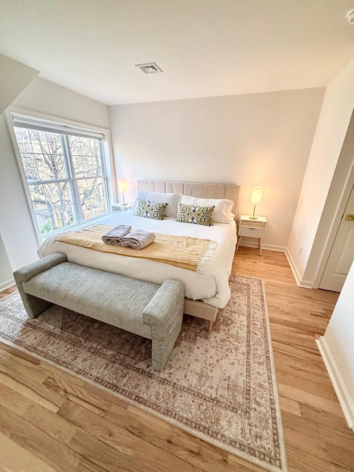 Une chambre à coucher avec un grand lit, un banc, une table de chevet et une fenêtre.