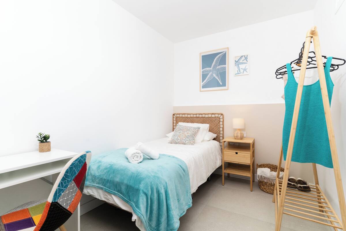 A bedroom with a bed, a desk, a chair, a hanging rack, and a small plant.