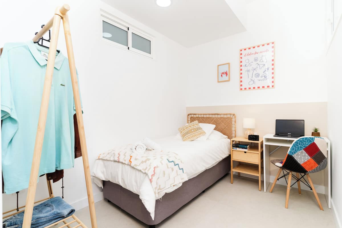 A bedroom with a bed, a desk with a laptop, a chair, a wooden hanger, and a window.