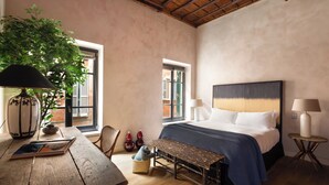 Un dormitorio con una cama grande, una mesa de madera, un banco, una planta en maceta y dos ventanas.