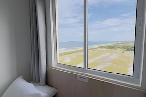 A room with a large window overlooking a beach and distant mountains. - Solarium Beach Club - Apartamento T2.904 (Itapoá)