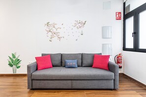 Ein graues Sofa mit roten und blauen Kissen, ein an der Wand befestigter Feuerlöscher und ein Fenster mit Jalousien.